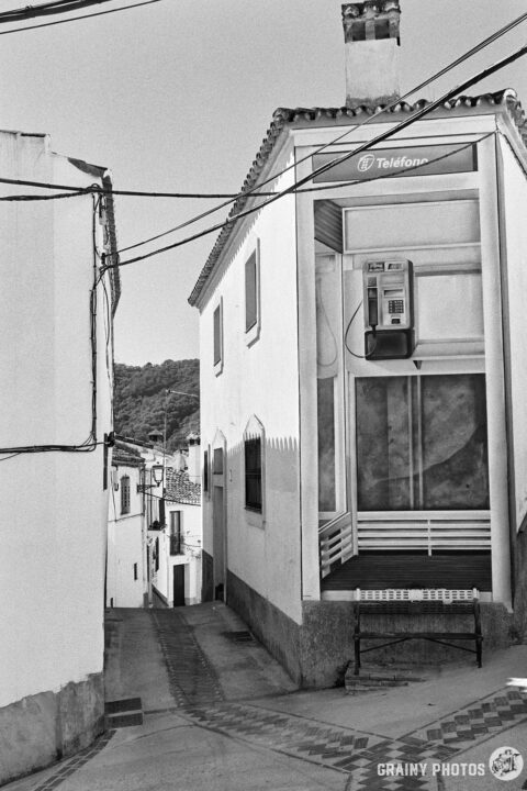 A black-and-white photo of a narrow street and white houses. On one house wall, there is a mural of a phone box, the full height of the house.