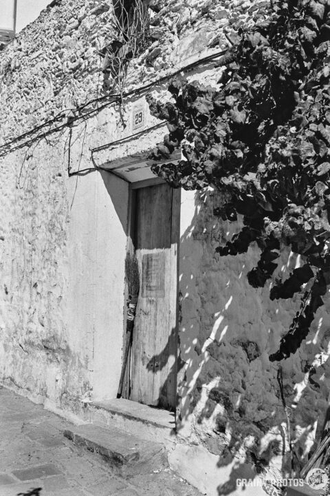 A black-and-white photo of an old white cottage with a wooden door. A broom is leaning against the door.