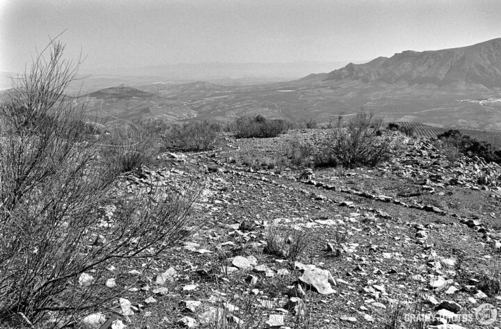 A rocky landscape with sparse vegetation, overlooking a vast valley and distant mountains in a black-and-white format. The terrain features a winding path, accentuated by various stones against a backdrop of rolling hills.