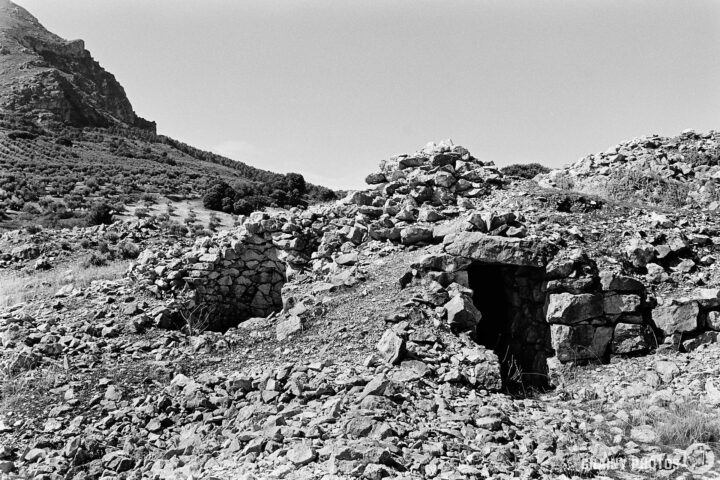 Ruins of an ancient stone structure at Los Caracoles, the entrance to a stonemason's hut, partially covered in rocks, set against a mountainous landscape. The area is dry, with sparse vegetation and a clear sky above, conveying a sense of history and nature's reclaiming of the land.