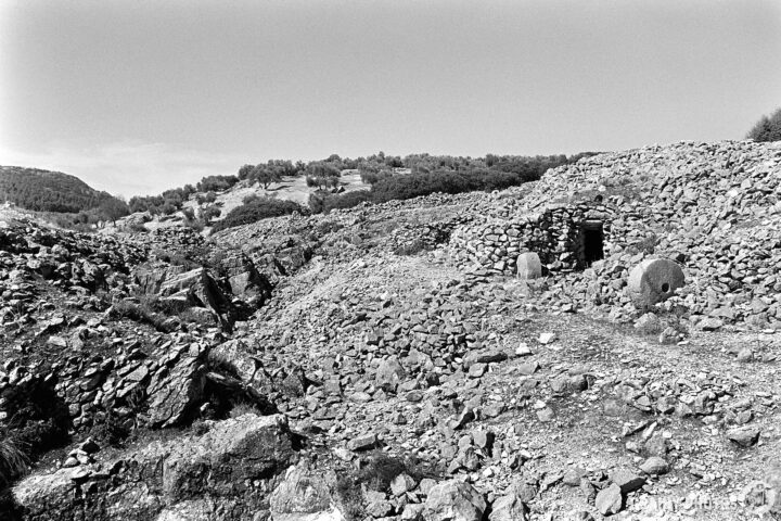 A black-and-white photo of a rock-strewn landscape with the entrance to a stone mason's hut. Either side of the entrance is a millstone - that is what they made here. A desolate landscape featuring rocky terrain, surrounded by greenery in the background.