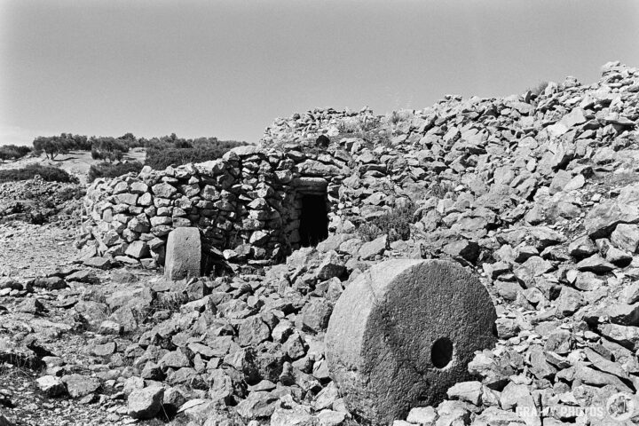 A desolate landscape featuring ancient stone ruins and large teo large millstones. The remnants of a structure are partially obscured by rocks, conveying a sense of history and abandonment under a clear sky.