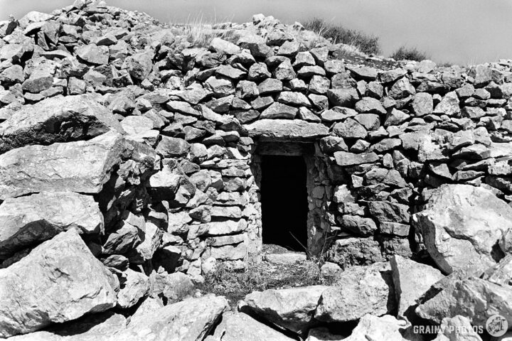A rustic, stone structure with a doorway, surrounded by large rocks and boulders, set against a clear sky. The image is in black and white, showcasing the textures of the stones and the natural landscape.
