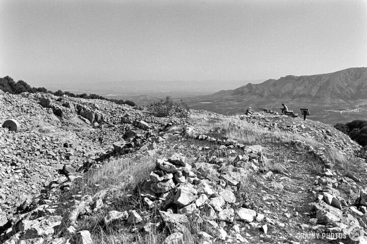 A panoramic view of ancient ruins on a hillside, with scattered rocks and vegetation, overlooking a vast valley and mountains in the distance. The scene is captured in black and white, emphasizing the historical significance of the landscape.