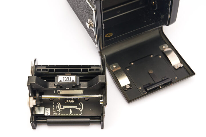An open Mamiya 645S camera with a removable film cartridge displayed beside it. The cartridge shows the number "120" and the word "JAPAN," highlighting the camera's design and film type.