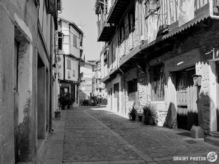 A narrow, cobblestone street lined with old stone houses, featuring wooden balconies and doorways, in a charming old town setting. Light casts shadows along the walls, creating a peaceful and nostalgic atmosphere.