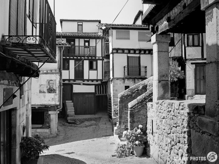 A street junction in Mogarraz, featuring traditional architecture with wooden balconies. The scene includes stone steps and a planter with flowers, set against a backdrop of historic buildings and a mural on the wall. Black and white photography.