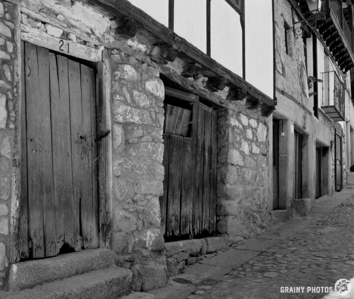 A black and white photograph of old stone buildings featuring wooden doors and a cobblestone pathway. The weathered façade exhibits rustic charm, with numbered door 21 visible and hints of historical architecture in a quiet alleyway.