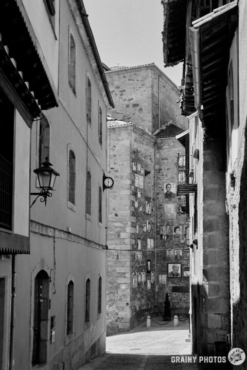 A narrow street in a Mogarraz features stone buildings with a clock and portraits (Las Caras de Mogarraz)on the church wall. The scene is captured in black and white, emphasizing the architectural details and the charming atmosphere of the area.