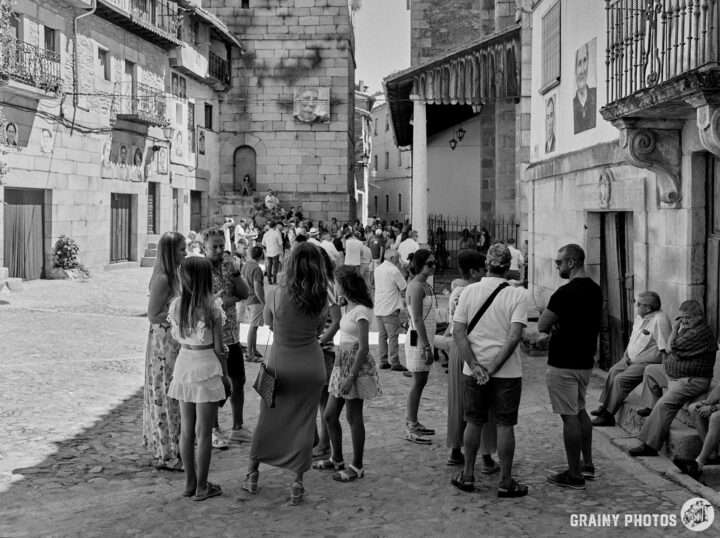 A lively town square scene in black and white, featuring groups of people engaging in conversation, with historic stone buildings and a prominent church tower in the background. Some individuals sit while others stand, immersed in the atmosphere.