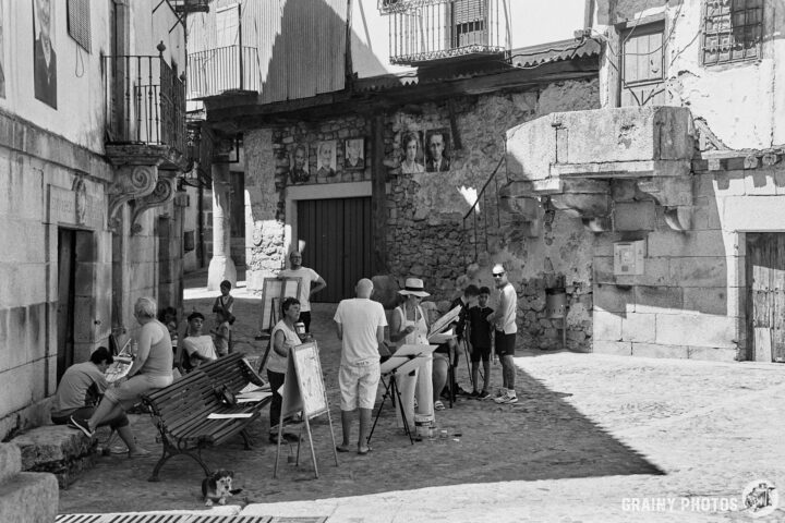 A group of artists gathered in a cobblestone square, some drawing or painting, while others observe. The scene features a mix of scenic architecture and rustic walls, capturing a lively, creative atmosphere in black and white.