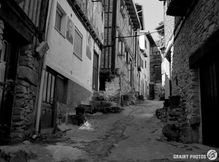 A narrow, winding street in Mogarraz, flanked by rustic stone buildings and wooden facades. The scene is captured in black and white, highlighting the texture of the cobblestone path and the architectural details of the structures.
