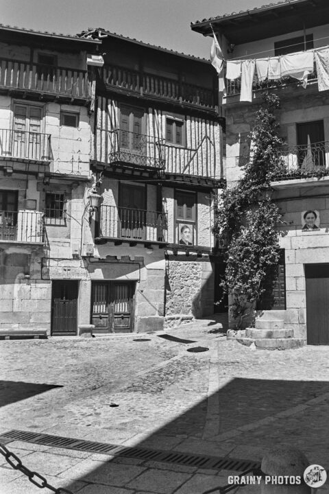 A small cobblestone plaza lined with historic buildings featuring balconies, a mix of stone and wood architecture, and greenery intertwining with a wall. The scene is illuminated by natural light, conveying a charming, rustic atmosphere.