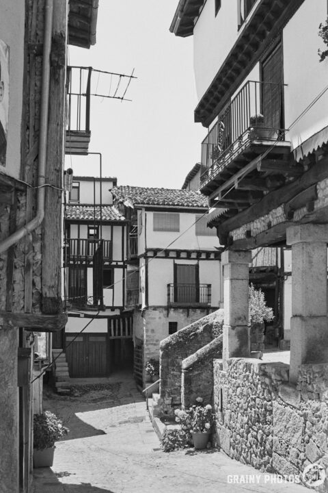 A narrow, cobblestone street lined with traditional buildings featuring balconies, stone walls, and flower pots. The scene captures the charm of a historic village, with a clear sky above and shadows cast by the overhanging structures.