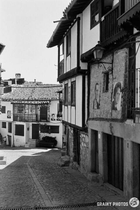 A cobblestone street lined with traditional buildings featuring wooden and stone facades. Some walls display black-and-white portraits (Las Caras de Mogarraz). The scene captures a quaint, historical ambiance in a rustic village setting.