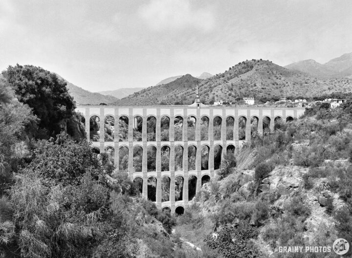 A black and white image of the scenic Acueducto del Águila spanning a valley, featuring numerous arches. The background showcases mountains and greenery, creating a dramatic landscape.