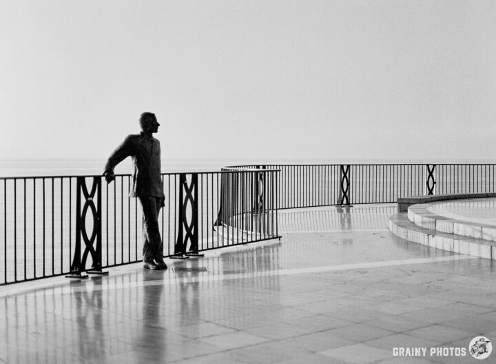 A solitary statue of King Alfonso XII by a railing looking out to sea, reflecting a calm atmosphere in black and white. The marble floor beneath adds to the serene setting and emphasizes the figure's contemplative pose.