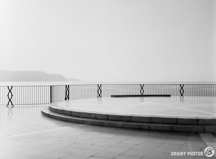 A misty seascape with a circular stone platform overlooking the water. The railing in the foreground features a geometric design, while distant hills fade into the hazy background, creating a serene and tranquil atmosphere.