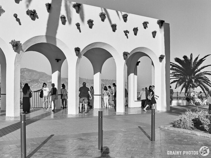 A scenic view of white arches, decorated with planters, on the Plaza Balcon De Europa. Groups of people enjoy the picturesque surroundings, with palm trees and mountains in the background, all captured in a black and white aesthetic.