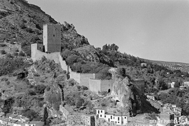 A historic stone fortress in Cazorla perched on a rocky hill, surrounded by trees and ruins. A medieval tower stands atop the structure, with remnants of walls flowing down the hillside, overlooking a small village below. The scene is captured in black and white.
