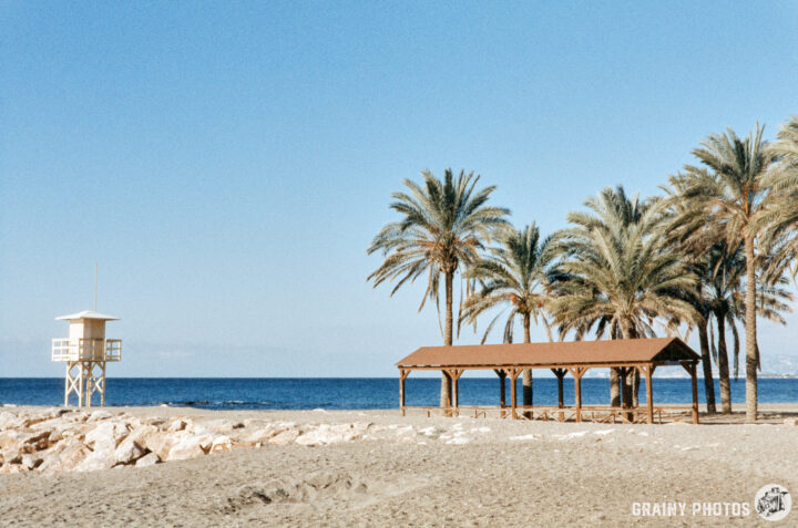 A quiet beach scene featuring a lifeguard tower, a shaded wooden pavilion, and palm trees against a clear blue sky, with gentle waves lapping at the shore.