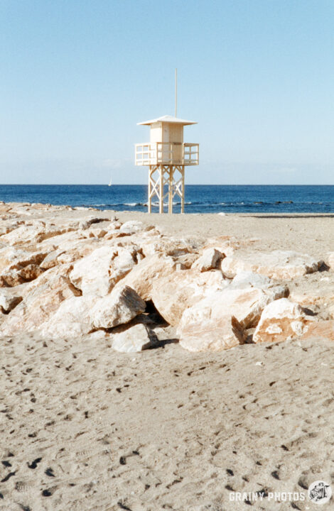 A lifeguard tower stands on a sandy beach beside large rocks, overlooking a calm blue sea under a clear sky.