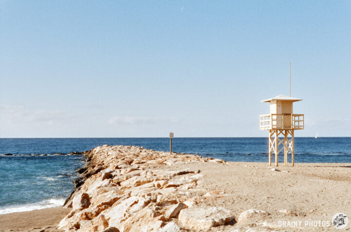 A solitary lifeguard tower stands on a rocky jetty by the beach, overlooking a calm ocean under a clear blue sky. The scene captures a serene coastal atmosphere with gentle waves lapping at the shore.
