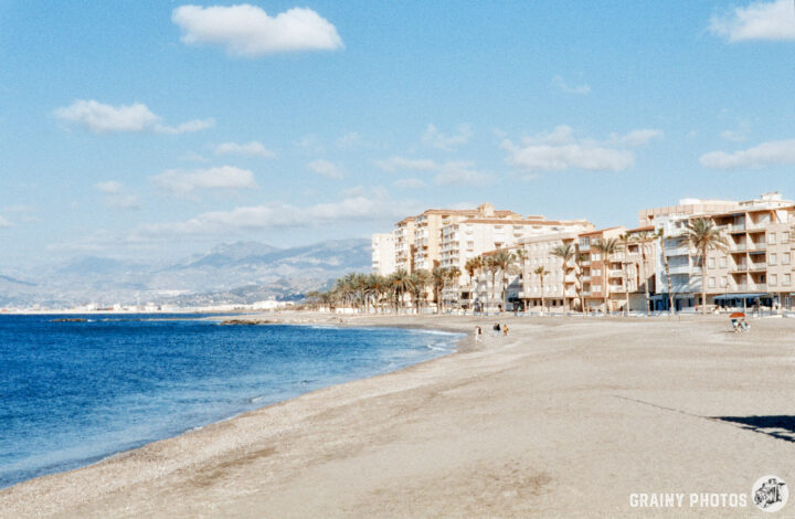 A sunny beach scene featuring a sandy shore, calm blue waters, and palm trees, with a background of seaside buildings and distant mountains under partly cloudy skies.