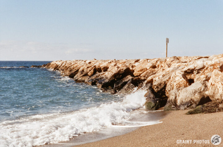 A rocky shoreline meets gentle waves along a sandy beach, with a distant post marking the coast. The ocean glistens under a clear sky, creating a tranquil seaside scene.