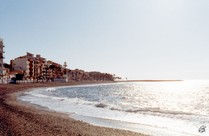 A serene beach scene at sunrise, featuring gentle waves lapping at the shore, pebbled coastline, and a view of low-rise buildings lined along the beach. The ocean sparkles under the early morning light, creating a peaceful atmosphere.