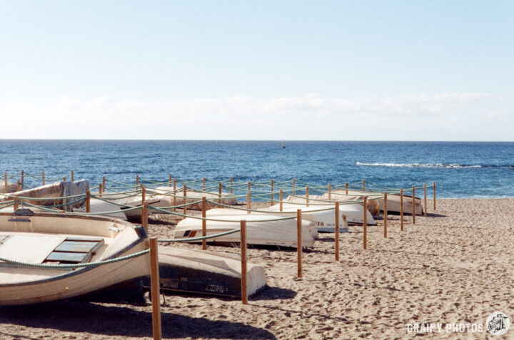A tranquil beach scene featuring several anchored boats along the shore, with gentle waves lapping at the sand and a clear blue sky in the background.