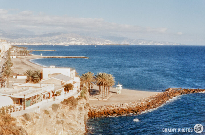 A scenic coastal view featuring a sandy beach, palm trees, and calm blue waters under a partly cloudy sky, with a rocky breakwater extending into the sea and buildings lining the shore in the distance.