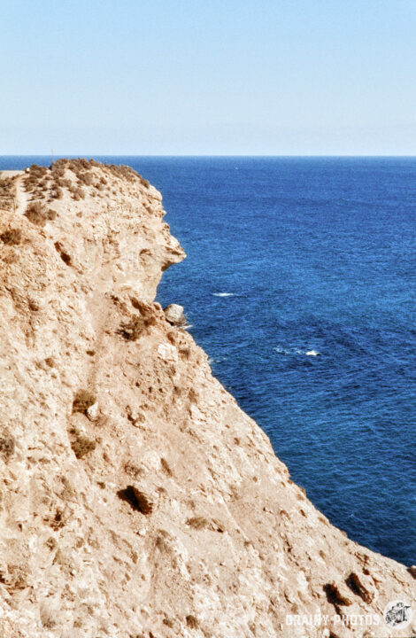 A rugged coastline featuring a steep cliff overlooking the calm blue ocean, with clear skies above and gentle waves lapping at the shore, showcasing the beauty of natural landscapes.