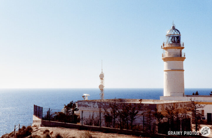 A coastal lighthouse stands atop a rocky outcrop, overlooking the Mediterranean. The structure features a classic tower design with a domed lantern room, surrounded by sparse vegetation and telecommunications antennas, against a clear blue sky.