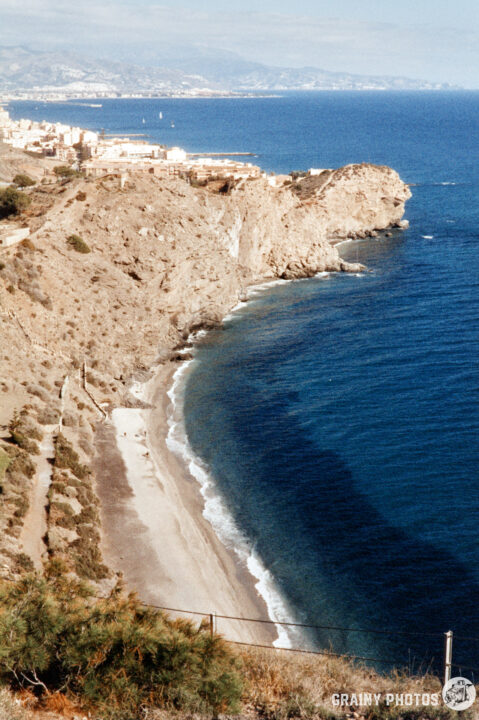 A coastal view showing a sandy beach along a rocky shoreline, with waves gently lapping at the shore. In the background, distant cliffs and a scenic town are visible under a bright sky.