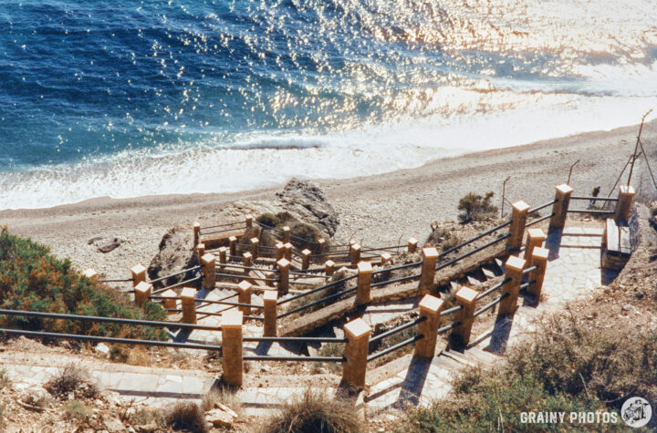 A winding staircase leads down to a sandy beach, with waves gently lapping at the shore. The scene showcases a sunlit coastline, framed by greenery and rocks, evoking a tranquil seaside atmosphere.