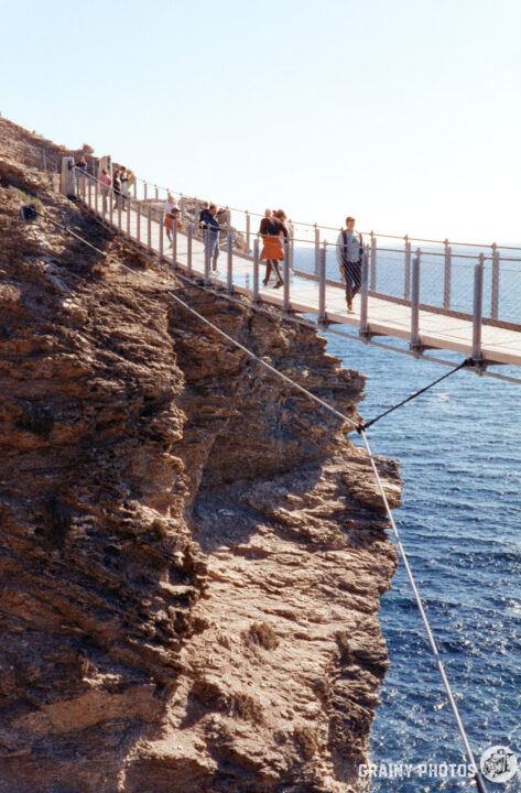 A scenic view of a suspension bridge stretching over a rocky cliff, with people walking across it. The ocean sparkles below, and the sky is clear, emphasizing the adventurous atmosphere of the location.