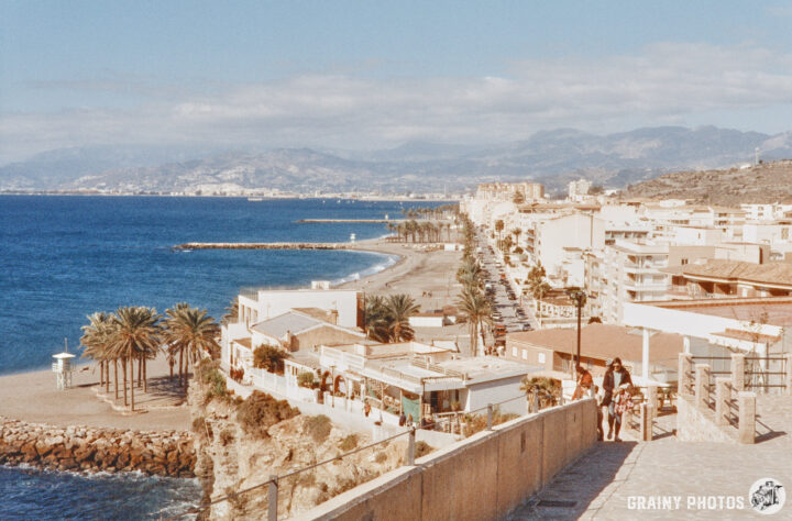 A scenic coastal view featuring a sandy beach lined with palm trees and buildings, set against a backdrop of mountains and a clear blue sky. The shoreline curves gently, and a promenade runs alongside with people walking.