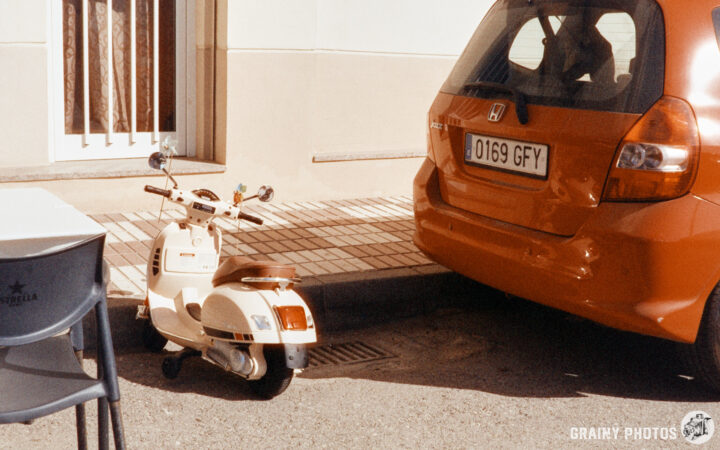 A vintage style mini cream scooter parked beside an orange car on a sunny street. The scene features a café table and chairs in the foreground, with a light-colored building in the background.
