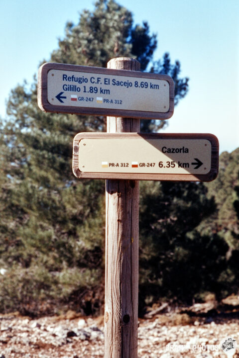 A wooden signpost in a natural setting displays distance markers for various locations: Refugio C.F. El Sacejo (8.69 km), Gillilo (1.89 km), and Cazorla (6.35 km), with directional arrows pointing towards each destination.