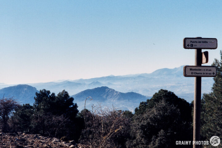 A scenic mountain view featuring signposts indicating directions to Puerto del Gaitero and Montesto. The landscape includes distant hills and trees under a clear blue sky.