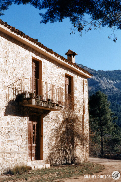 A rustic stone house features a balcony, surrounded by trees and mountains under a clear blue sky, showcasing a serene natural landscape.