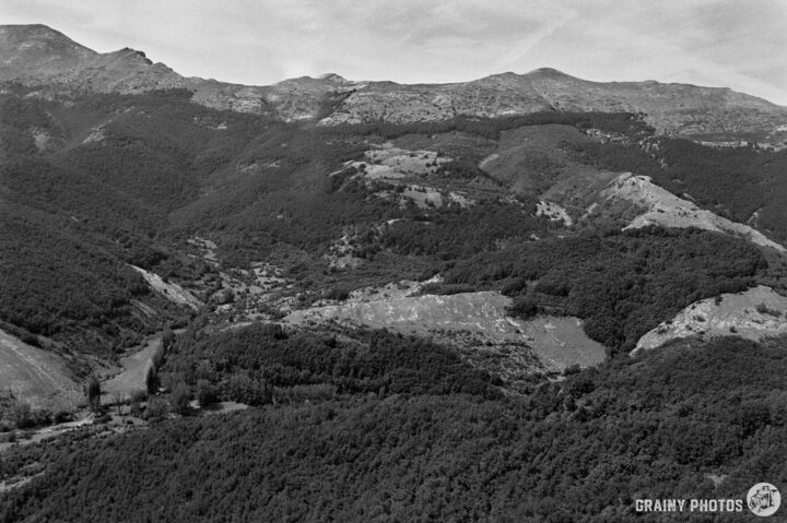 A black and white aerial view of a mountainous landscape featuring rolling hills, dense forests, and a meandering river cutting through the valley. The scene captures the natural beauty and rugged terrain of the area.