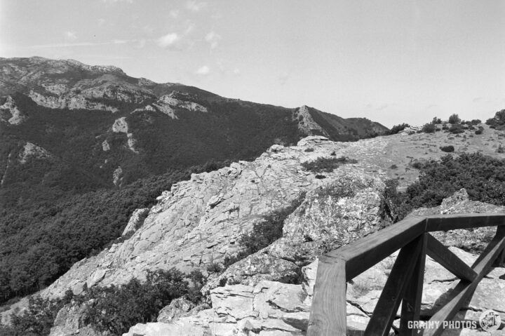 A viewing platform with a wooden railing overlooks a mountainous landscape. The scene is captured in black and white, highlighting the textures of the rocks and foliage against the distant peaks.