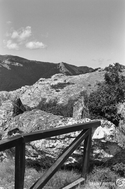 A scenic black-and-white landscape featuring rocky terrain and distant mountains, with a wooden fence on the viewing platform framing the foreground. Fluffy clouds drift in a clear sky above the natural vista.