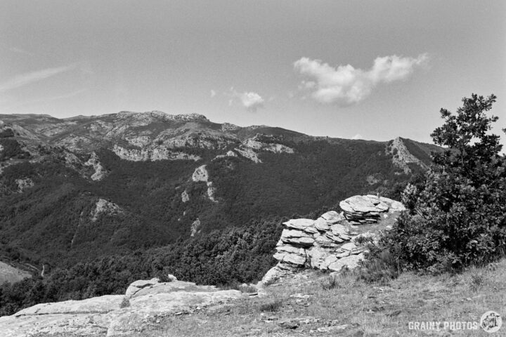 A black-and-white photograph showcasing a rocky outcrop in the foreground with a vast mountain landscape in the background, featuring rolling hills and a partially cloudy sky. The scene conveys a serene atmosphere of nature.