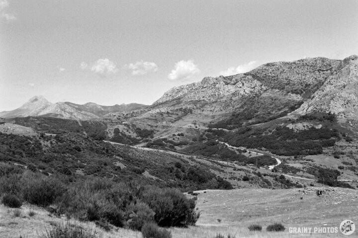 A black and white landscape featuring rolling hills and a winding road. Rocky mountains rise in the background, surrounded by patches of greenery and sparse vegetation under a clear sky with a few clouds.