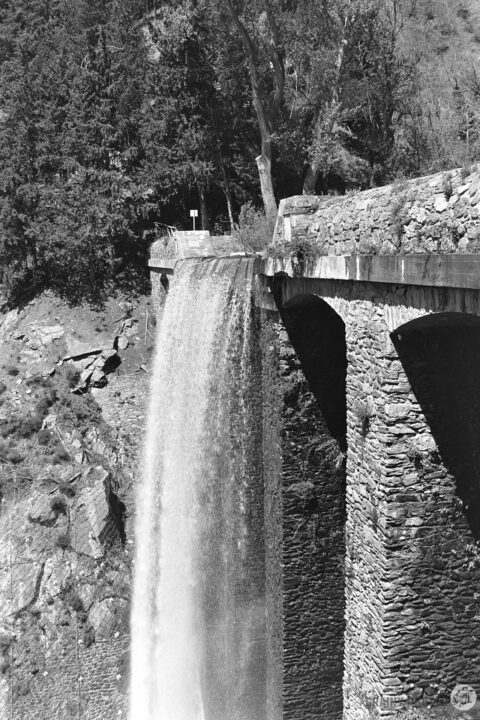 A black and white photograph of a waterfall cascading over a stone dam, surrounded by trees and rocky terrain, capturing the natural beauty and engineering of the structure.