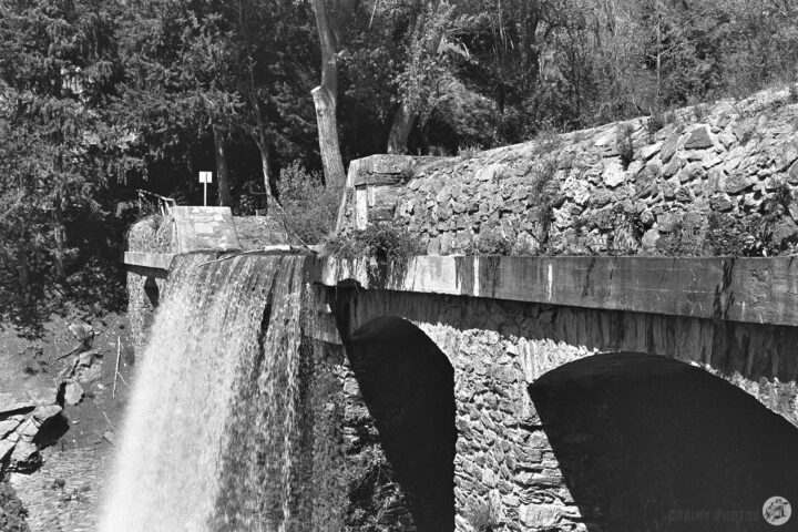 A stone dam with a waterfall cascading down. Lush greenery surrounds the area, creating a serene natural landscape. The image is presented in black and white, emphasizing texture and contrast.