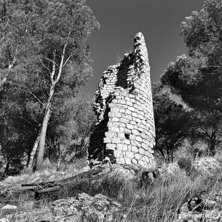 A weathered stone tower stands amidst trees, surrounded by rocky terrain. The structure, partially collapsed, showcases ancient architecture, blending with the natural landscape in a monochrome setting.